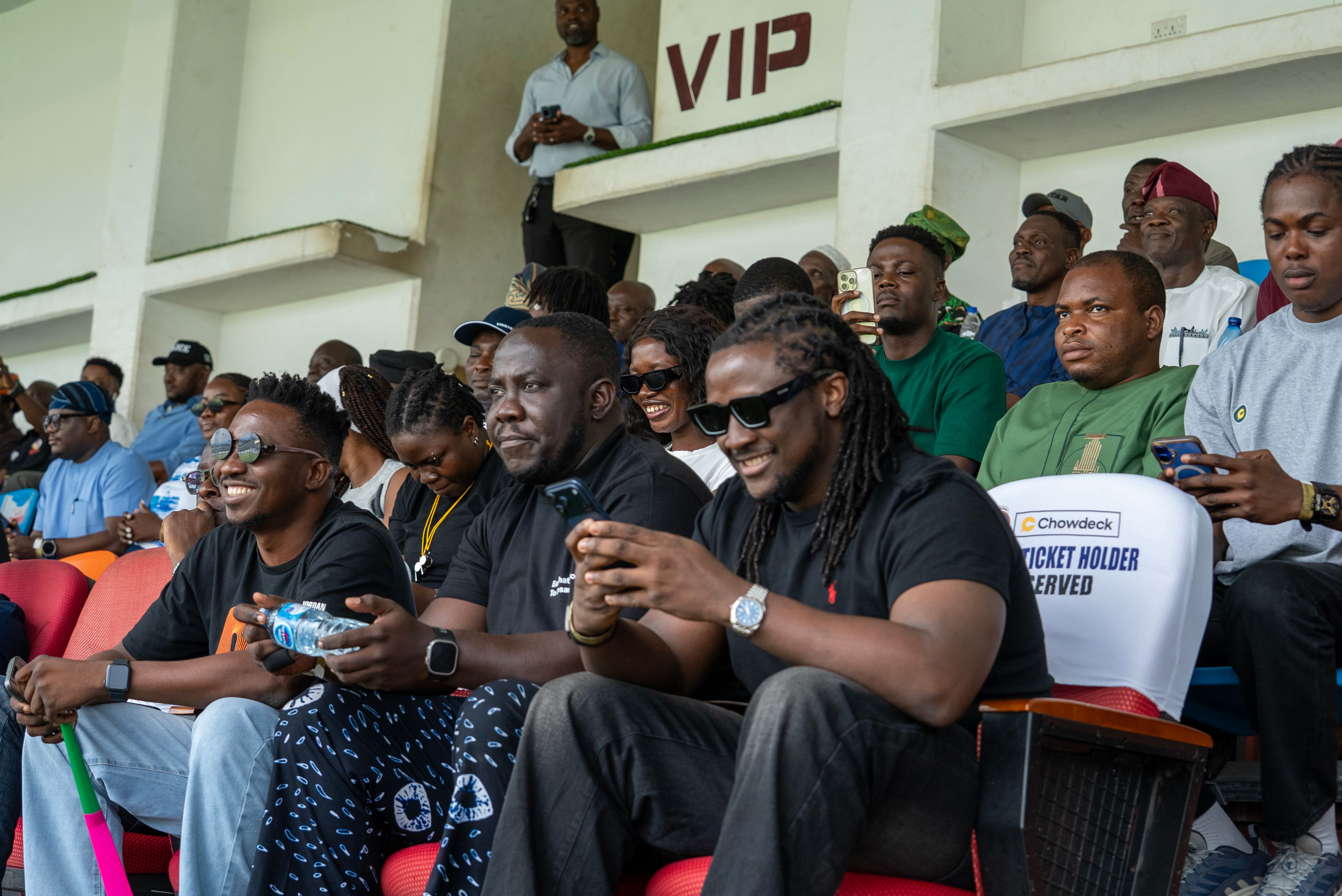 Spectators sit in a stadium stand watching a football match.