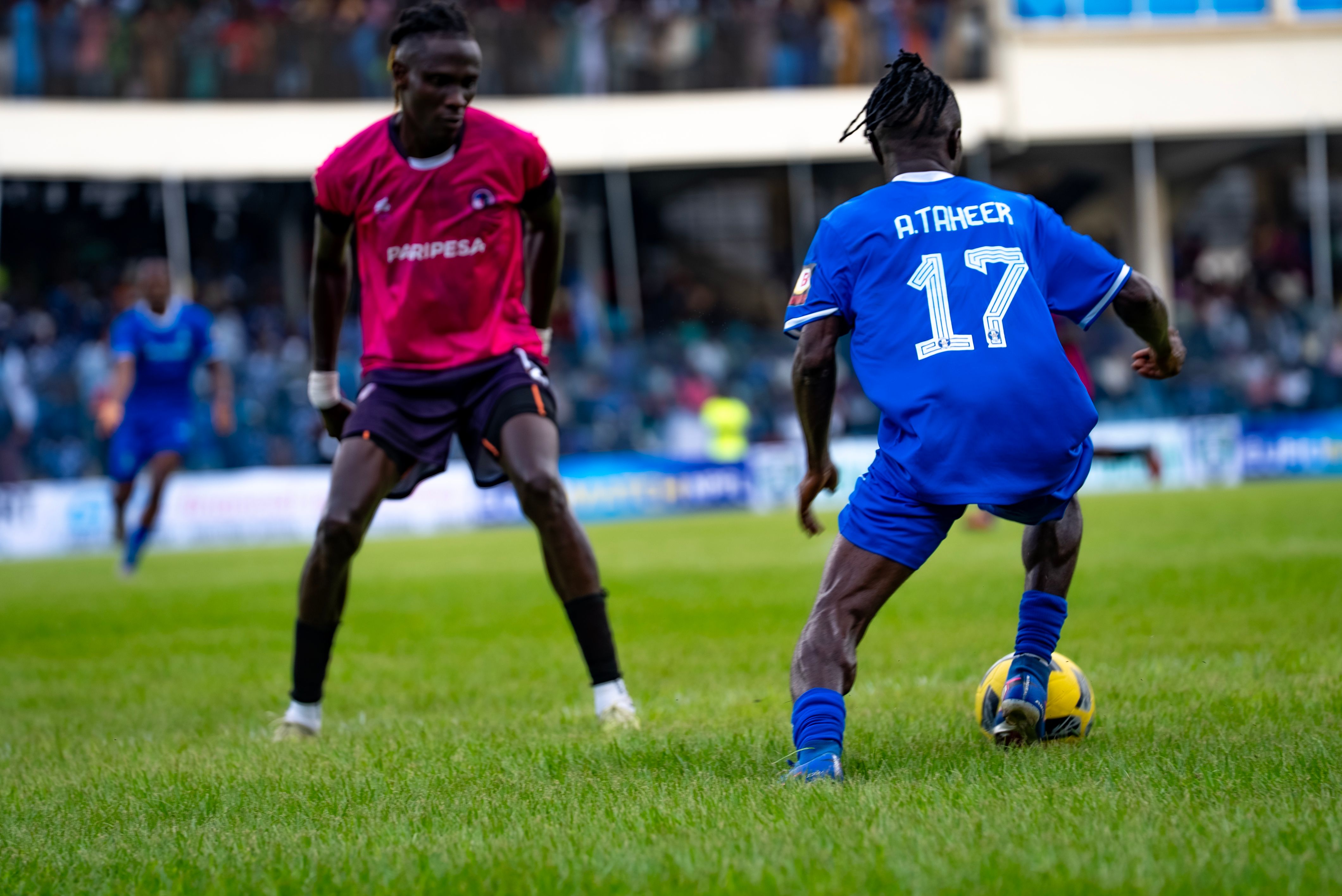 A Shooting Stars player in a blue kit, number 17 and named A. Taheer, dribbles the ball while a defender from Interlagos in a pink kit closes in, during a football match in front of a full stadium crowd.