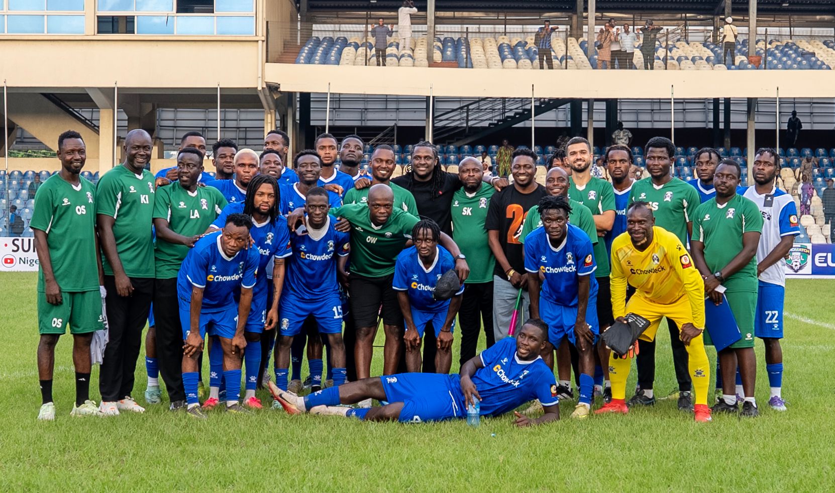A football team group photo on the pitch after a match: players in blue kits sponsored by Chowdeck stand alongside coaching staff in green tracksuits, smiling and posing together, with one player lying on the ground in a playful pose in front of the group